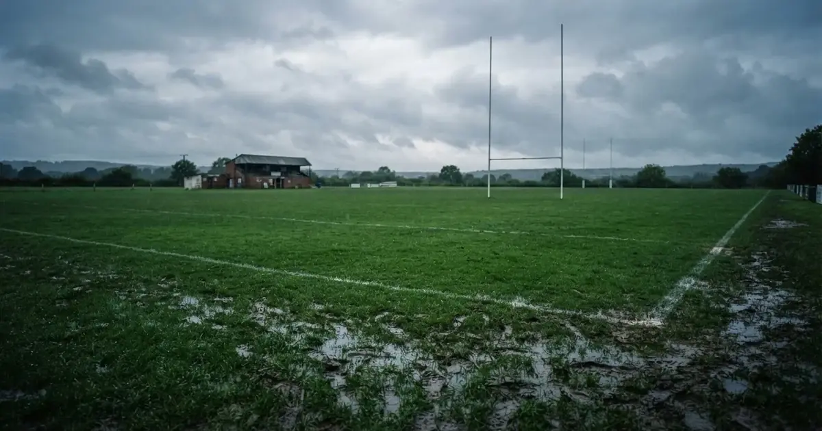 Boisko do rugby ze słupami bramki w oddali pod pochmurnym niebem, mokra trawa i ślady butów.