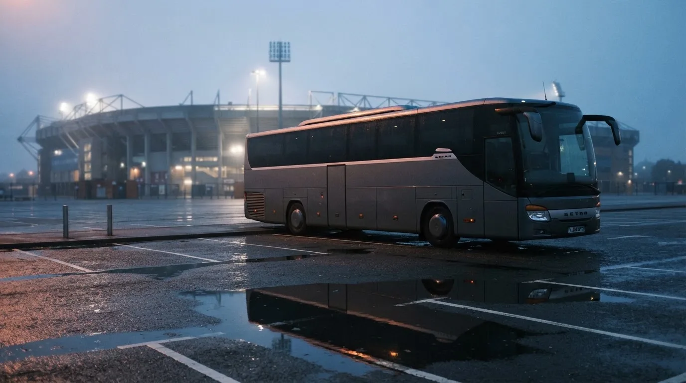 Autokar stoi na parkingu przed stadionem o świcie, rozmyte kontury trybun w tle, pusty plac i mokry asfalt.